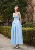 Woman wearing a light blue strapless gown with corset detailing and a full skirt, standing outdoors.