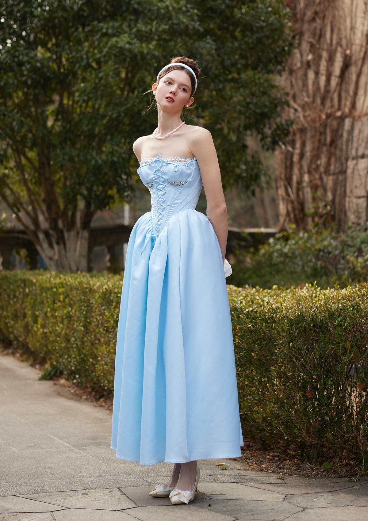 Woman wearing a light blue strapless gown with corset detailing and a full skirt, standing outdoors.
