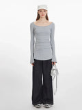 Woman wearing a grey long-sleeved ribbed knit top with a scoop neckline, ruched side detail, flared cuffs, and a side slit hem, paired with dark wash wide-leg jeans and a silver handbag.