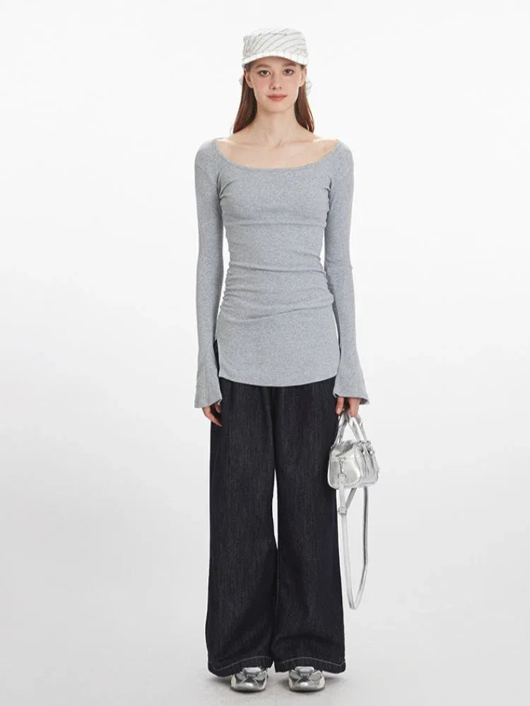 Woman wearing a grey long-sleeved ribbed knit top with a scoop neckline, ruched side detail, flared cuffs, and a side slit hem, paired with dark wash wide-leg jeans and a silver handbag.
