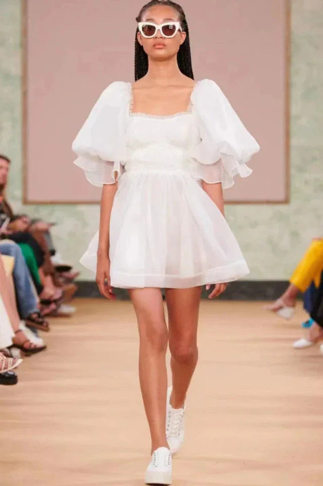Model wearing a white organza mini dress with puffy sleeves and a square neckline on a runway
