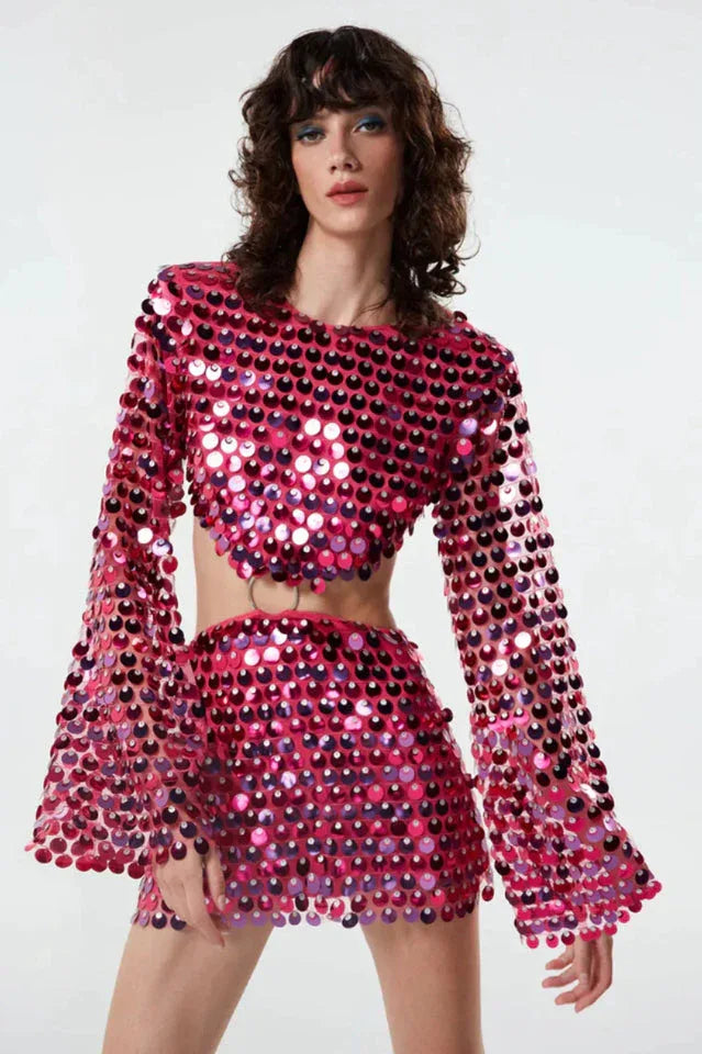 Woman wearing a pink sequined long-sleeve cropped top with cut-out detail and ring hardware, paired with a matching pink sequined mini skirt. Main image