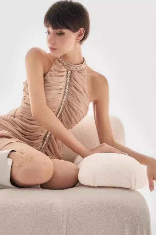 Woman posing on a chair wearing a ruched halter neck mini dress with embellished neckline in a light beige color.
