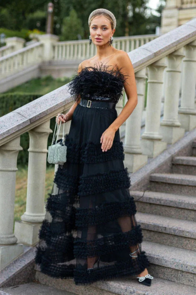 Woman wearing a black strapless evening gown with feather and sequin details and a tiered ruffle skirt, holding a silver clutch, standing on stairs outdoors.