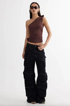 Woman wearing a brown one-shoulder ruched top and dark wash wide-leg cargo pants, posing with hands on hips.