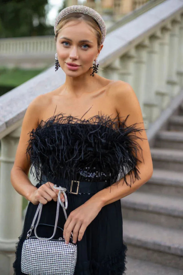 Close-up of a woman in a black strapless evening gown with a feather-trimmed bodice and sequin band, holding a silver clutch.