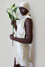 woman wearing a cream-colored satin mini dress with a halter neckline and a long sash, holding lilies