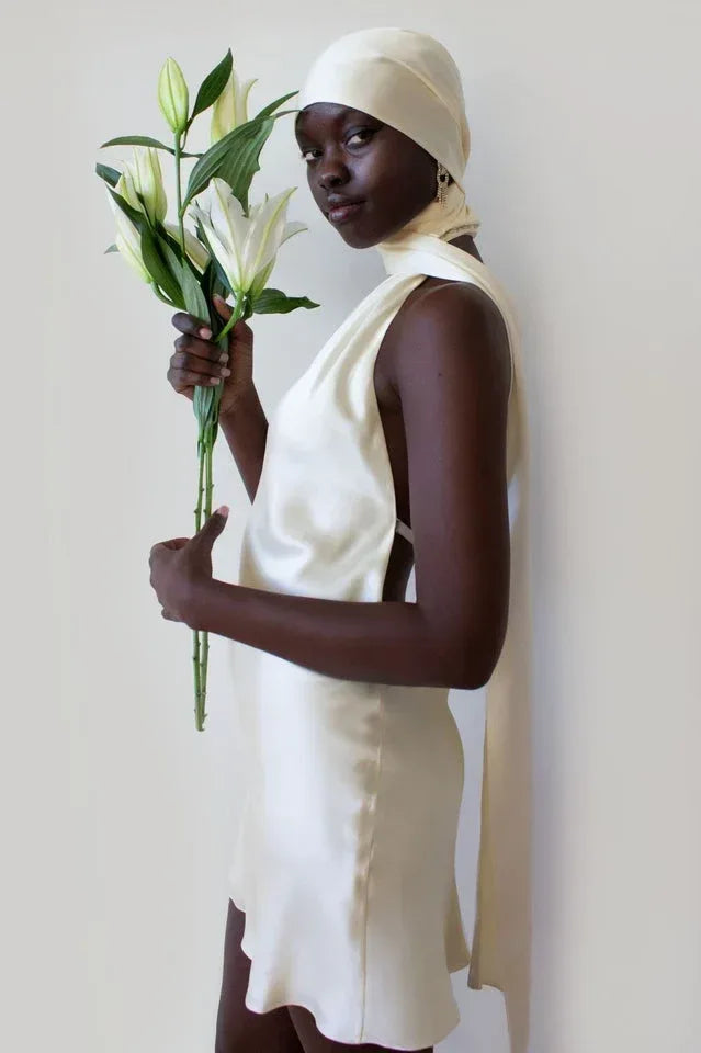 woman wearing a cream-colored satin mini dress with a halter neckline and a long sash, holding lilies