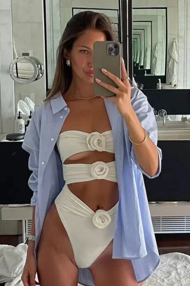 Woman taking a mirror selfie in a white strapless swimsuit with cut-out detailing and floral appliques, wearing an open blue shirt.