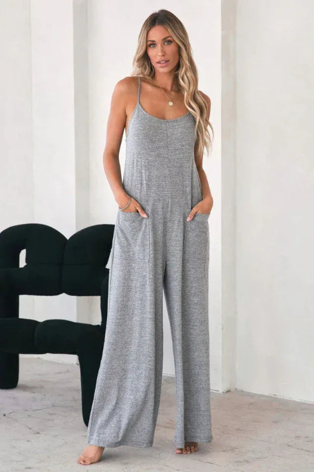 Woman in a grey jersey knit wide leg jumpsuit with patch pockets and spaghetti straps, standing.