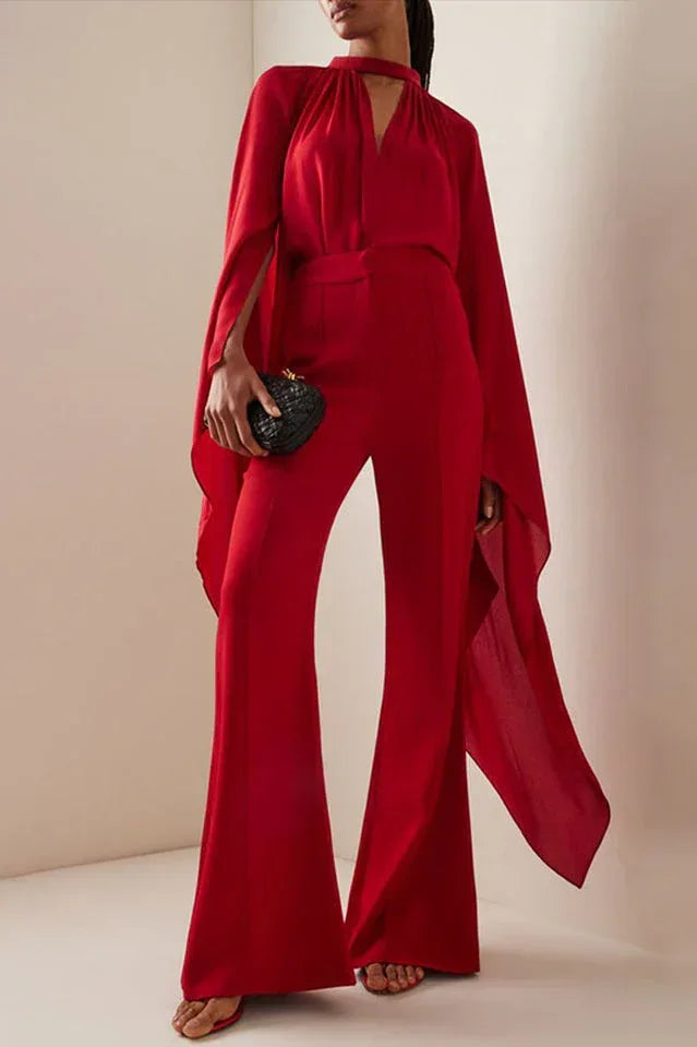 Woman wearing a red crepe wide leg jumpsuit with draped cape sleeves and a halter neckline.
