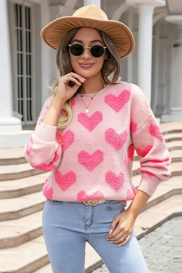 Woman wearing a pink heart pattern sweater, fedora hat, and sunglasses.
