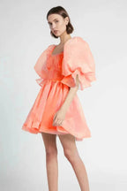 Woman wearing a coral organza mini dress with puffy sleeves and a square neckline, shown from the side