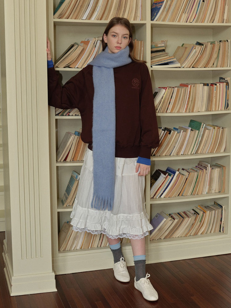 Woman wearing a brown velour sweatshirt with a light blue scarf, white ruffled skirt, grey socks, and white sneakers, standing in front of a bookshelf.