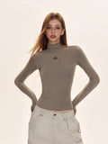 Woman wearing a taupe turtleneck top with a black embroidered cross logo on the chest.