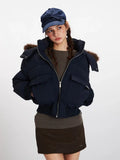 Woman wearing a navy blue cropped puffer jacket with a fur-trimmed hood, dark brown skirt, and brown boots, with hands in pockets.