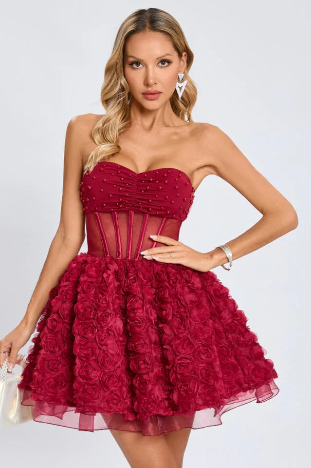 Woman wearing a short, strapless red dress with a ruched bodice adorned with small beads, a sheer corset waist, and a full skirt covered in fabric roses, presented against a white background.