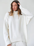 Woman adjusting the hem of a white oversized turtleneck sweater while wearing light wash wide-leg jeans.