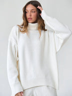 Woman adjusting the hem of a white oversized turtleneck sweater while wearing light wash wide-leg jeans.