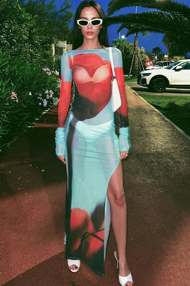Woman wearing a long, sheer, light blue dress with an abstract red and black floral print, long sleeves, and a high slit, accessorized with white sunglasses and a white handbag.
