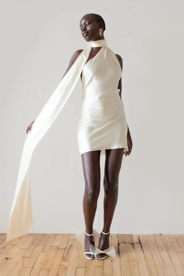 woman wearing a cream-colored satin mini dress with a halter neckline and a long sash, standing with one arm outstretched