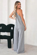 Back view of a woman in a grey jersey knit wide leg jumpsuit with patch pockets and spaghetti straps.