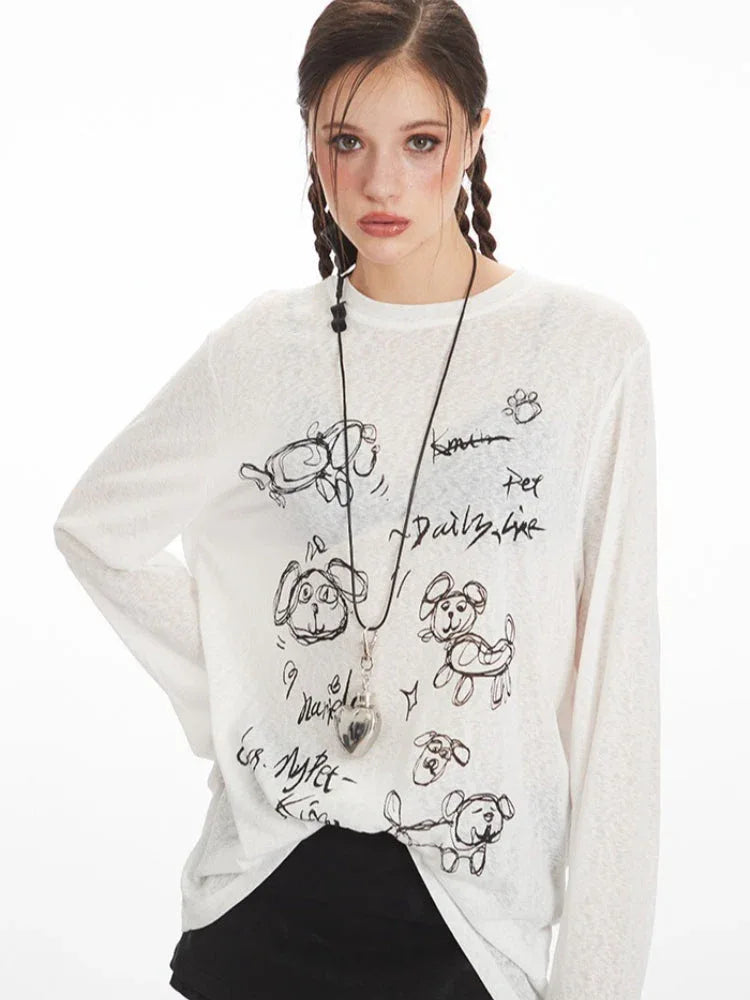 Front view of a woman wearing a white long-sleeve sheer knit t-shirt with a black dog graphic print and text, and a silver heart pendant necklace.