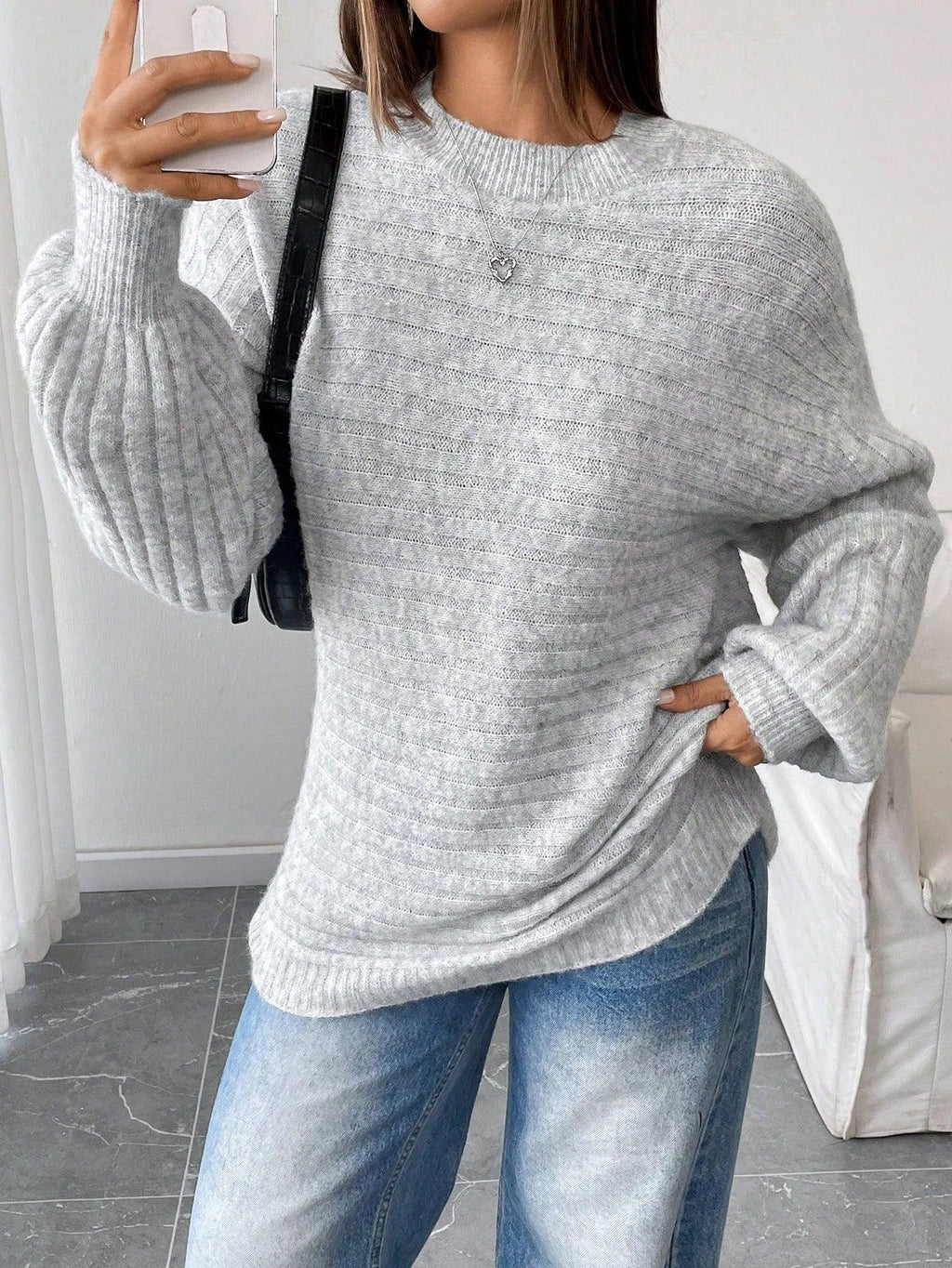 Woman taking a selfie while wearing a light grey oversized knit sweater and blue denim jeans, accessorized with a black handbag.