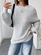 Woman taking a selfie while wearing a light grey oversized knit sweater and blue denim jeans, accessorized with a black handbag.