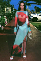 Woman standing on a pathway wearing a long, sheer, light blue dress with an abstract red and black floral print, long sleeves, and a high slit, holding sunglasses.
