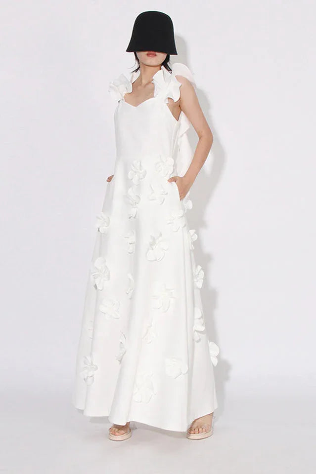 Woman wearing a white floor-length A-line dress with 3D floral appliqués and ruffled shoulder straps, showing side pockets, standing in front of a white background.