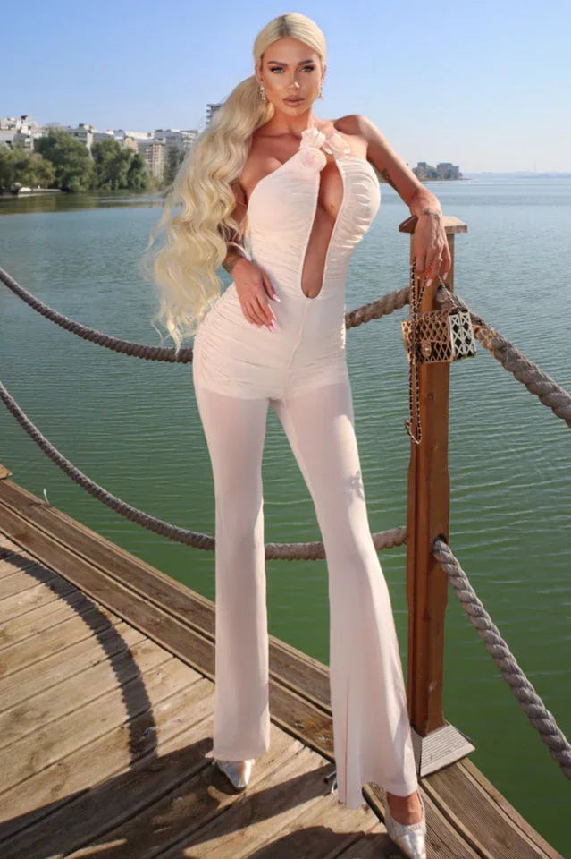 Woman wearing a light pink ruched jumpsuit with a plunging neckline and floral details, standing on a wooden deck by the water.