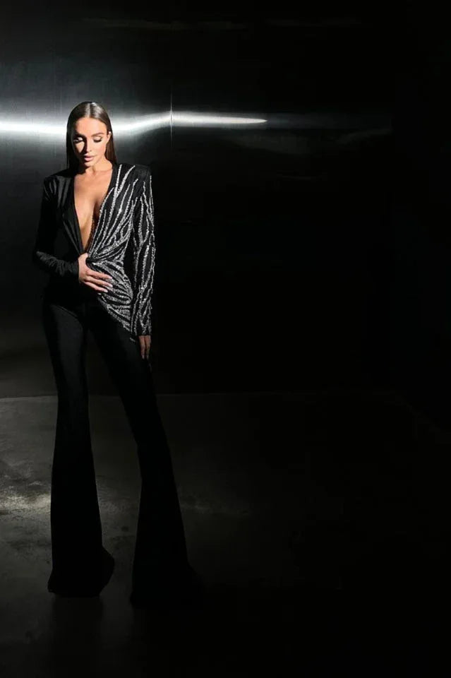 Woman posing in a black jumpsuit with a deep V-neck and flared legs in a dark setting.