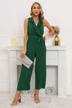 Woman wearing a green sleeveless pleated jumpsuit with a ruffled V-neck and a tie belt.