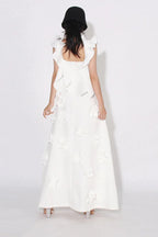 Back view of a woman wearing a white floor-length A-line dress with 3D floral appliqués and ruffled shoulder straps, standing in front of a white background.
