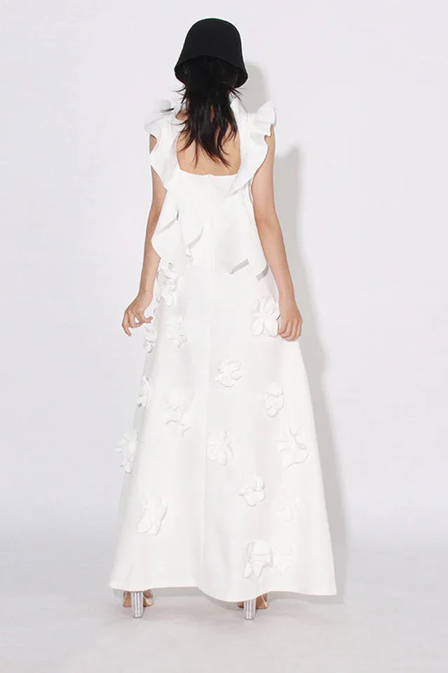 Back view of a woman wearing a white floor-length A-line dress with 3D floral appliqués and ruffled shoulder straps, standing in front of a white background.