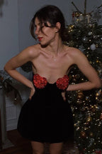 Woman in black strapless velvet mini dress with red heart embellishments on bust, standing in front of a Christmas tree.