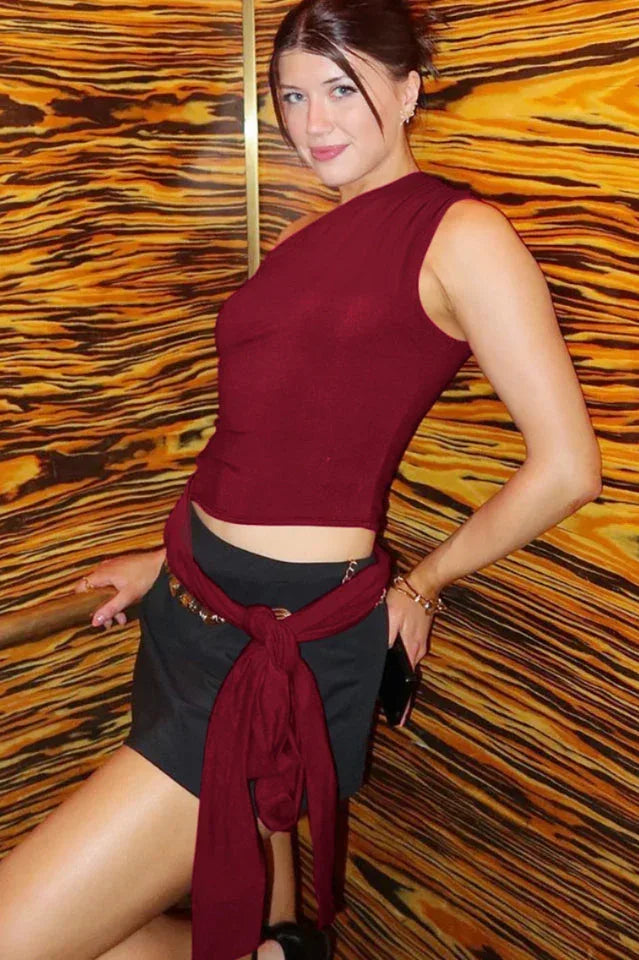 Back view of a woman wearing a burgundy one-shoulder draped top with light blue textured shorts.