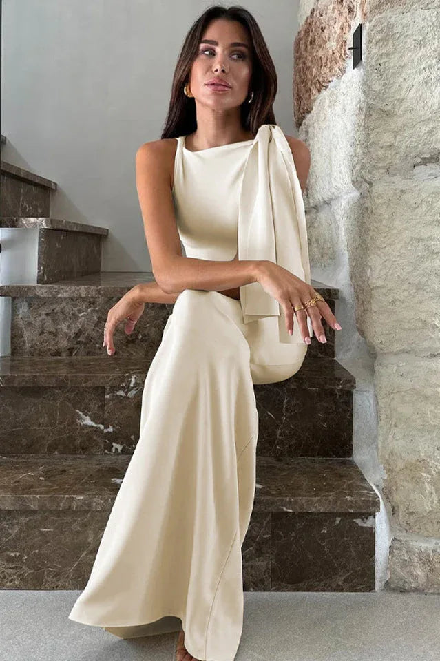 Woman sitting on stairs wearing a cream sleeveless column dress with an asymmetrical neckline and draped shoulder detail.