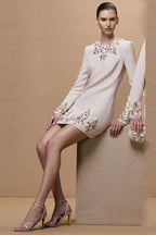 Full body shot of a woman in a cream long-sleeve mini dress with crystal and pearl embellishments, paired with silver heels.