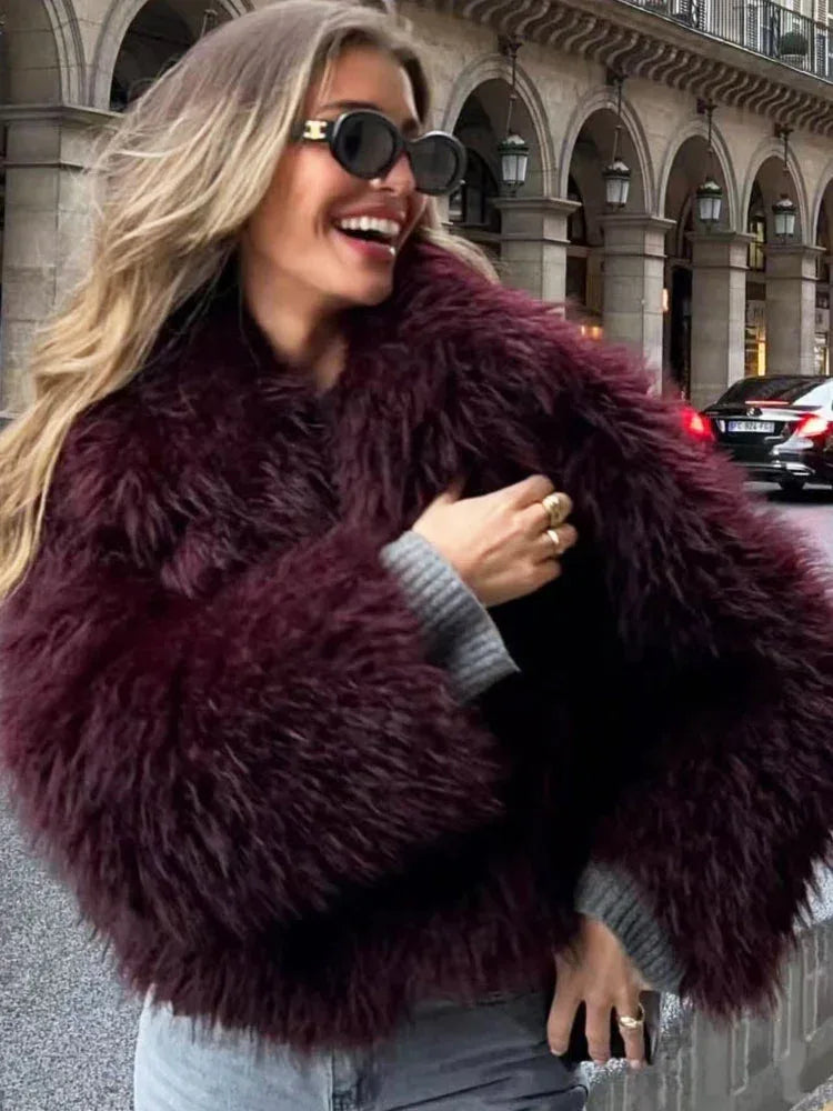 Woman wearing a burgundy shaggy faux fur cropped jacket and grey sweater, with gold rings, smiling.