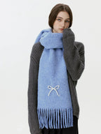 Woman wearing a light blue scarf with a white bow embellishment and fringe, hands tucked into sweater.