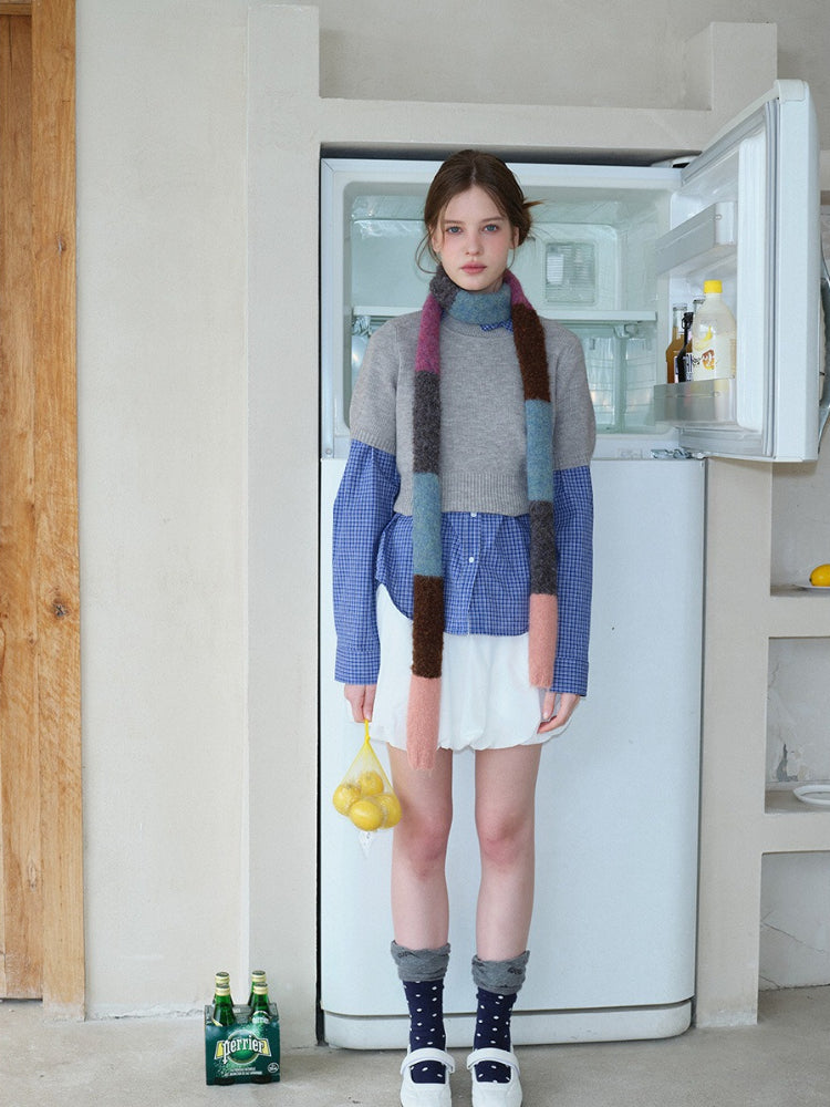 Model in a layered outfit with a grey sweater, blue gingham shirt, white skirt, and a vibrant scarf.
