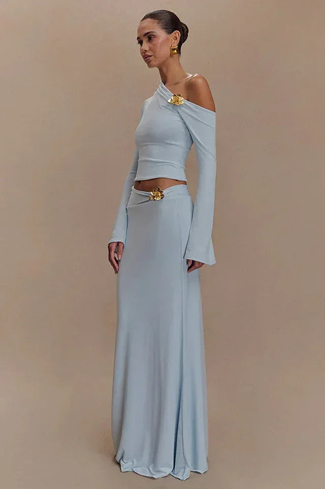 Woman wearing a light blue long-sleeved cropped top and a floor-length light blue skirt with a floral embellishment on the shoulder and hip.