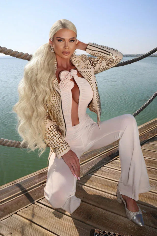 Woman in a light pink ruched jumpsuit with a plunging neckline and floral details, kneeling on a wooden deck.