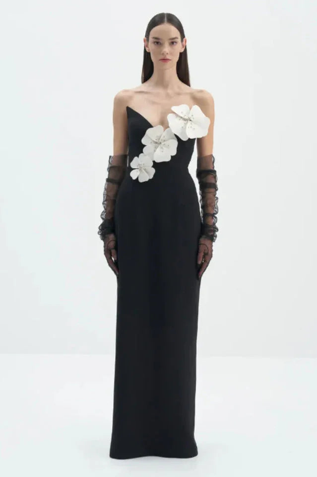 Woman wearing a black column gown with a sweetheart neckline and white floral applique on the bodice, paired with black sheer ruffled gloves. Main image