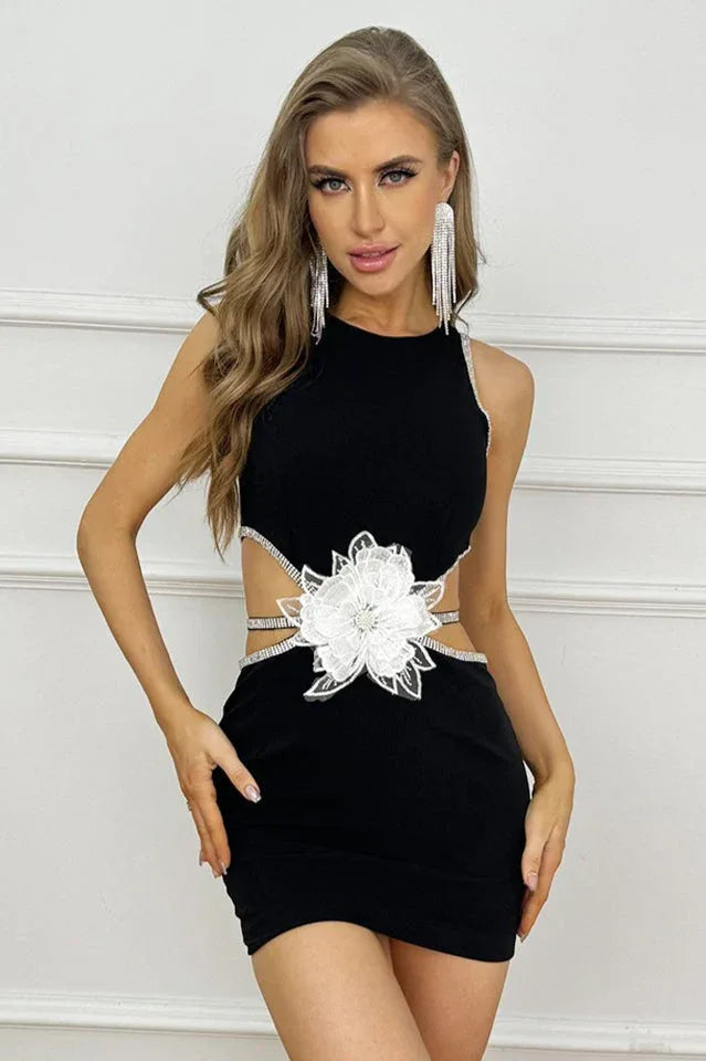 Woman wearing a black sleeveless bodycon mini dress with side cutouts, embellished trim, and a large white floral applique at the waist.