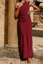 Woman posing in a burgundy satin draped dress with a one-shoulder design.
