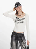 Woman wearing a white long-sleeve graphic t-shirt with a scoop neckline and studded details, looking to the side.