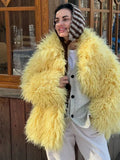 Woman wearing a yellow shaggy faux fur coat over a grey cardigan and white t-shirt, with a striped beanie.